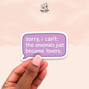 May include: A purple sticker with white border, held by a hand, features the text "sorry, i can't. the enemies just became lovers." The sticker has a speech bubble shape. The background is a light pink color.