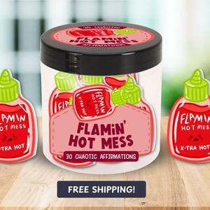 Puede incluir: Un tarro de 30 afirmaciones caóticas "Flamin' Hot Mess". El tarro es blanco con una tapa negra y presenta una botella de salsa picante roja y blanca de dibujos animados con el texto "Flamin' Hot Mess" y "X-Tra Hot".