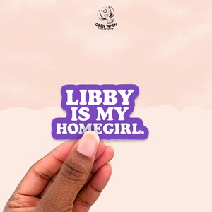 May include: A purple sticker with the text "LIBBY IS MY HOMEGIRL" in white, held by a hand. The sticker has a rounded rectangle shape. The background is a light peach color.