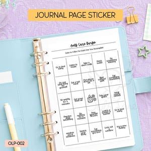 May include: A light blue binder with a "Self Care Bingo" journal page sticker. The sticker features a grid of self-care activities. The text "Journal Page Sticker" is at the top. A yellow pen and a gold paper clip are also visible.