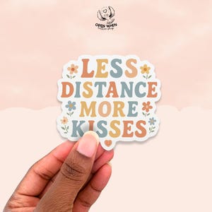 May include: A sticker with the text "LESS DISTANCE MORE KISSES" in a retro font, surrounded by floral accents. The sticker has a white border and is held by a hand against a peach-colored background. The sticker is from the "Open When Letter Shop".