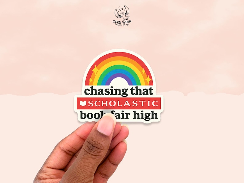 Puede incluir: Una pegatina con un dise&ntilde;o de arco&iacute;ris y el texto "chasing that SCHOLASTIC book fair high". La pegatina tiene un fondo blanco y un borde rojo. La pegatina est&aacute; sujeta por una mano.