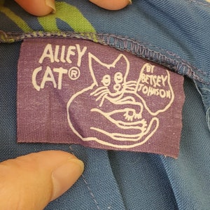 Può includere: Etichetta in tessuto viola con testo bianco che recita "Alley Cat® by Betsey Johnson". L'etichetta presenta un gatto dei cartoni animati con occhi grandi e una mano che tiene un gatto piccolo.