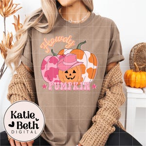 May include: A brown t-shirt with a graphic of three pumpkins, one wearing a pink cowboy hat. The pumpkins are pink and orange with a cow print design. The text reads "Howdy Pumpkin" in pink and orange.
