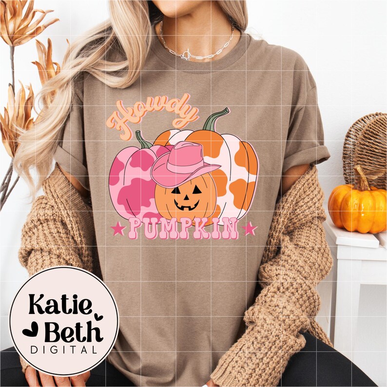 May include: A brown t-shirt with a graphic of three pumpkins, one wearing a pink cowboy hat. The pumpkins are pink and orange with a cow print design. The text reads "Howdy Pumpkin" in pink and orange.