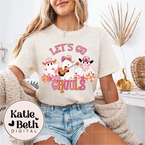 May include: Cream-colored t-shirt with the text "LET'S GO GHOULS" in pink and orange. The graphic features three cartoon ghosts wearing cowboy hats and bandanas, with one playing a guitar. The shirt is part of a Halloween-themed collection.