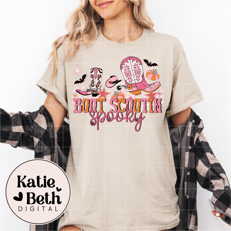 May include: Beige t-shirt with a colorful graphic featuring cowboy boots, a hat, stars, bats, and a pumpkin. The text "Boot Scootin' Spooky" is displayed in pink and orange. The shirt has short sleeves.