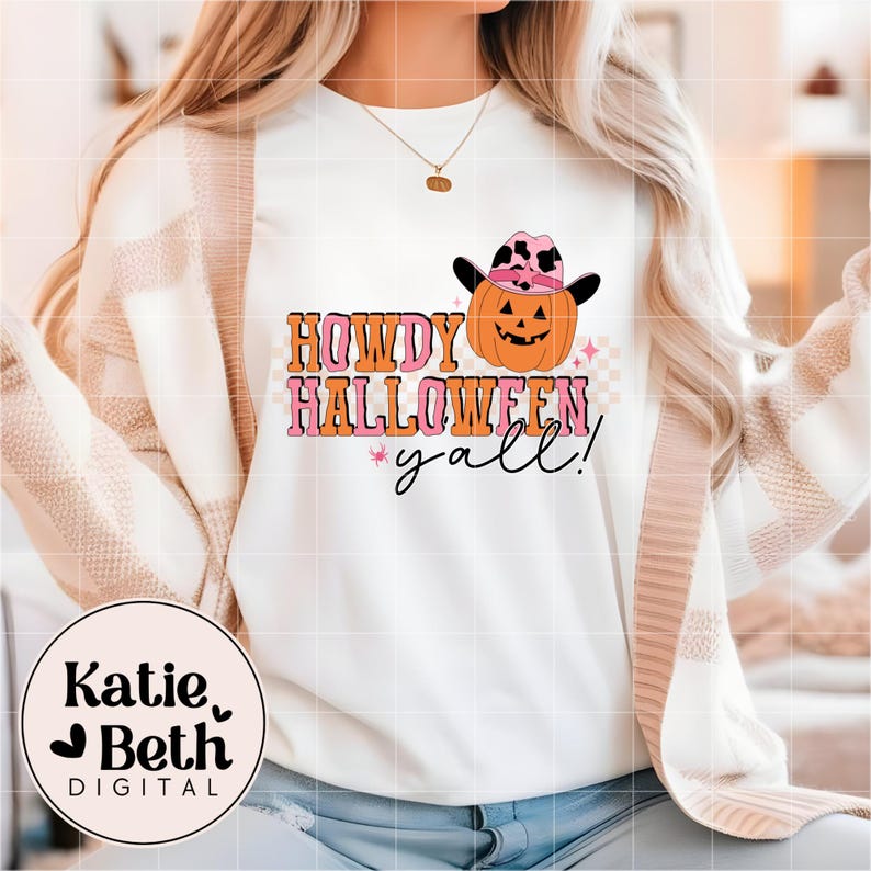 May include: White sweatshirt with a graphic that says "Howdy Halloween y'all!" in orange and pink lettering. The design includes a smiling pumpkin wearing a pink and black cowboy hat. A pink star and a checkered pattern are also included.