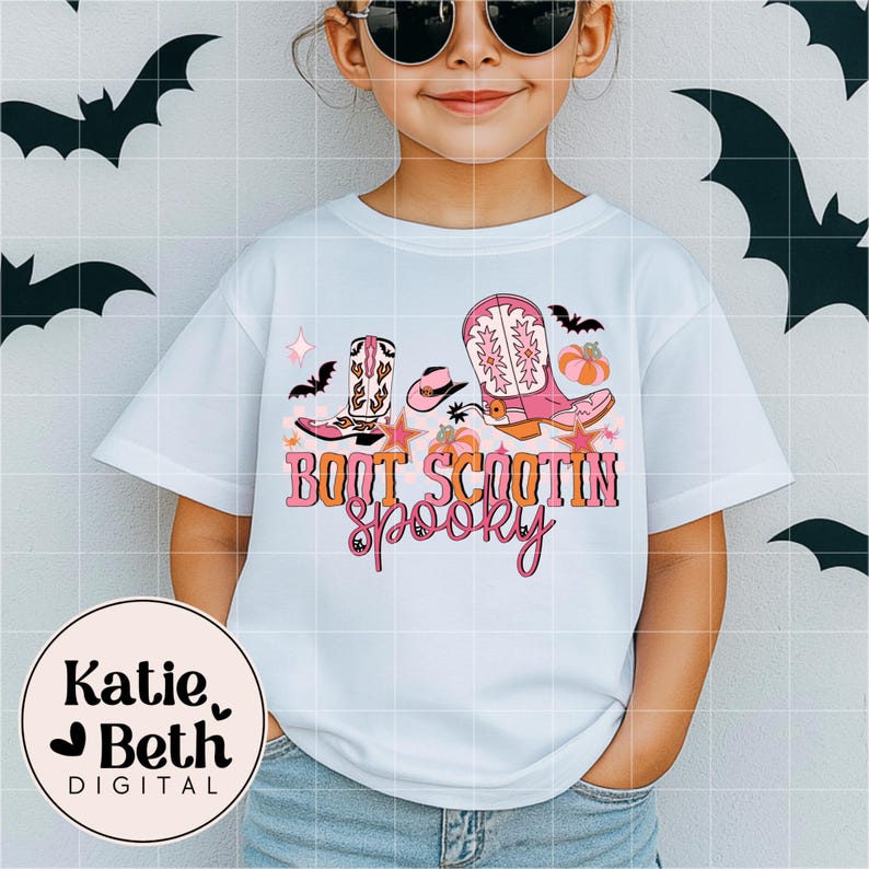 May include: Light blue t-shirt featuring a colorful graphic with cowboy boots, a hat, bats, and pumpkins. The text "Boot Scootin' Spooky" is displayed in pink and orange. A circular logo with the text "Katie Beth Digital" is in the lower left.