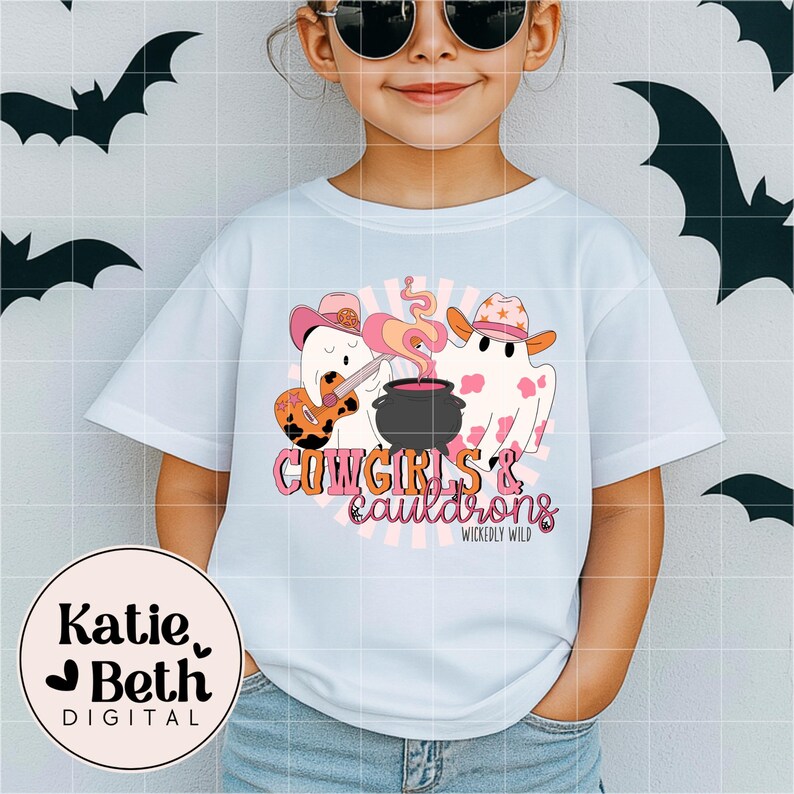 May include: A light blue t-shirt featuring a Halloween-themed graphic. The design includes two ghost figures wearing cowboy hats, one playing a guitar, and a cauldron with pink smoke. The text reads "Cowgirls & Cauldrons" and "Wickedly Wild".