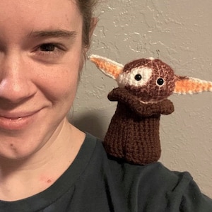 May include: A person wearing a dark blue shirt with a brown crocheted toy on their shoulder. The toy has large white eyes and orange ears.