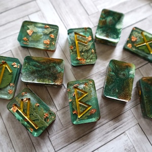 May include: Ten green and gold resin dice with gold runes and dried botanicals inside. The dice are square and have rounded edges.