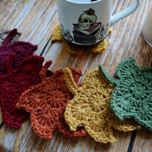 Puede incluir: Un juego de seis posavasos de ganchillo con forma de hoja en colores verde, amarillo, naranja, rojo, rojo oscuro y gris. Cada posavasos tiene forma de hoja de arce con un tallo. Una taza de cerámica blanca con un gráfico de calavera descansa.