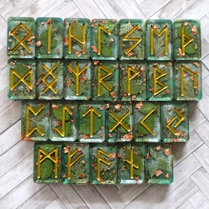 May include: Set of 24 green and gold resin rune tiles with gold leaf accents. Each tile features a different rune symbol.