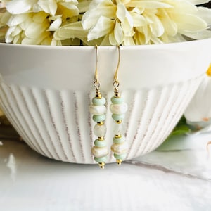 Op de afbeelding: Goudkleurige oorhangers met lichtgroene, witte en gouden kralen. De oorbellen hebben een gelaagd ontwerp met een mix van ronde en onregelmatig gevormde kralen, wat een unieke en elegante uitstraling creëert. Gepresenteerd voor een witte kom.