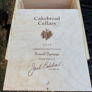 May include: A light-colored wooden wine box with the text "Cakebread Cellars" and "2018 Cabernet Sauvignon Suscol Springs Napa Valley". The box has a sliding lid and a small, round wooden knob.