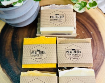 Puede incluir: Cuatro barras de jabón natural hecho a mano con etiquetas de papel marrón. Las etiquetas tienen un patrón de panal y el texto "CHAUTAUQUA SOAP CO." Los aromas del jabón son Prado de primavera, Naranja, Pomelo y Naranja, y Romero y Naranja.