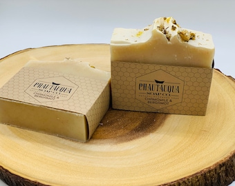 May include: Two bars of natural handmade soap with a brown paper label that reads "CHAUTAUQUA SOAP CO. CHAMOMILE & BERGAMOT". The soap is on a wooden surface.