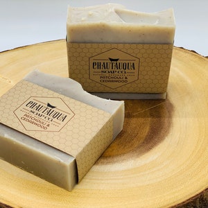 Op de afbeelding: Twee repen natuurlijke handgemaakte zeep met een bruin papieren etiket waarop staat "Chautauqua Soap Co. Natuurlijke handgemaakte zeep Patchouli & cederhout". De zeep ligt op een houten oppervlak.