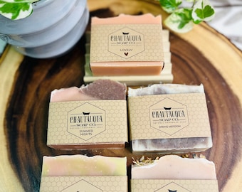 Puede incluir: Cuatro barras de jabón natural hecho a mano con diferentes aromas. Las barras de jabón están envueltas en papel marrón con una etiqueta blanca que dice "Chautauqua Soap Co." y el aroma de cada barra. Los aromas son "Lovely", "Summer Nights", "Spring Meadow" y "Secret Garden".