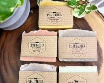 Puede incluir: Cuatro barras de jabón natural hecho a mano envueltas en papel marrón con un patrón hexagonal. Las barras de jabón están etiquetadas con el texto "CHAUTAUQUA SOAP CO." y las fragancias: "Romero & Naranja", "Árbol de Té", "Patchouli & Cedro" y "Lavanda".