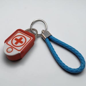 Puede incluir: Un llavero con un dije rectangular rojo y blanco que presenta un símbolo de cruz médica y un pequeño acento circular. El dije está unido a una anilla de metal plateado conectada a un lazo de cordón trenzado de color azul claro.