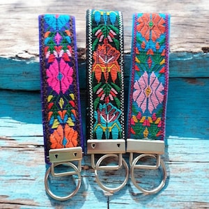 May include: Three colorful keychains with embroidered floral patterns. Each keychain has a silver metal ring and a silver metal clasp. The keychains are displayed on a blue wooden surface.