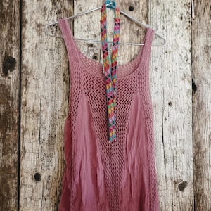 May include: A dusty rose-colored tank top with a crochet overlay, hanging on a silver hanger. A colorful, patterned lanyard is draped over the hanger. The background is a weathered, light-colored wooden surface.