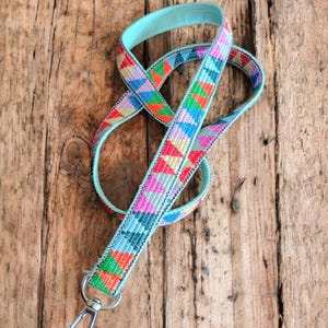 May include: A colorful lanyard with a geometric pattern in red, pink, green, blue, and yellow. The lanyard has a light blue backing and a silver metal clasp. The lanyard is laying on a wooden surface.