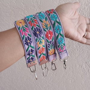 May include: Four lavender fabric wristlet keychains featuring vibrant embroidered floral patterns in shades of yellow, red, purple, pink, and teal. Each strap is finished with a silver-toned metal clasp and a circular split ring for keys.