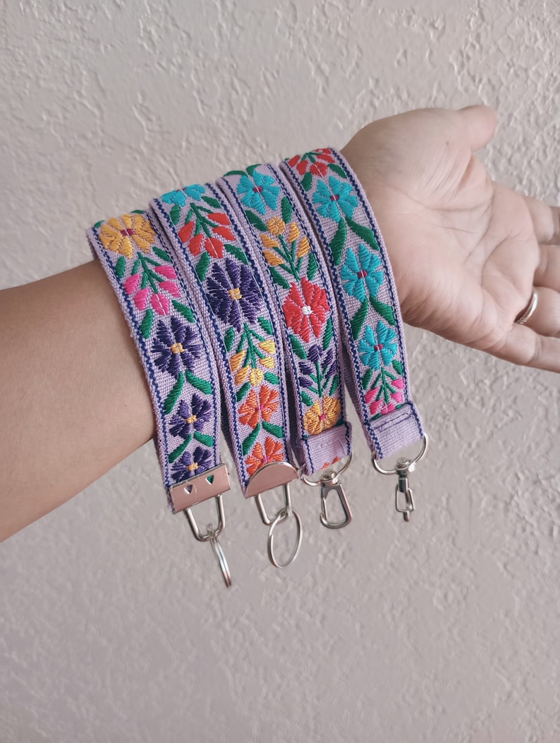 May include: Four lavender fabric wristlet keychains featuring vibrant embroidered floral patterns in shades of yellow, red, purple, pink, and teal. Each strap is finished with a silver-toned metal clasp and a circular split ring for keys.
