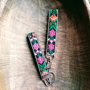 May include: Two black key fobs with floral embroidery in pink, orange, and green. The key fobs have silver-colored metal clasps and rings. The key fobs are displayed on a wooden surface.
