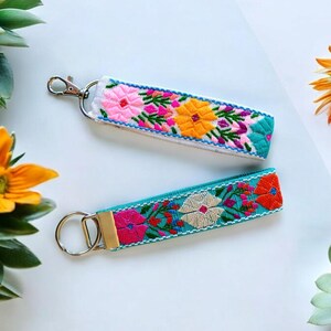 May include: Two colorful embroidered keychains with silver key rings. The keychain on the top is white with pink, yellow, and green embroidery. The keychain on the bottom is turquoise with pink, white, and orange embroidery.