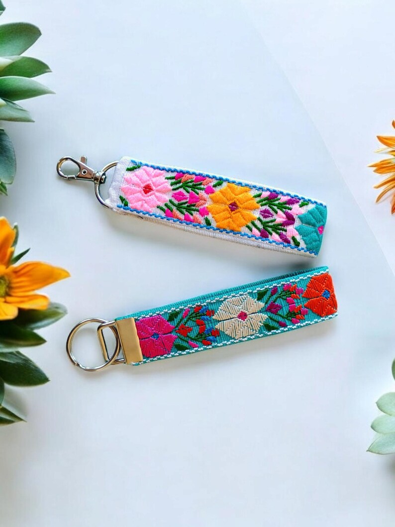 May include: Two colorful embroidered keychains with silver key rings. The keychain on the top is white with pink, yellow, and green embroidery. The keychain on the bottom is turquoise with pink, white, and orange embroidery.