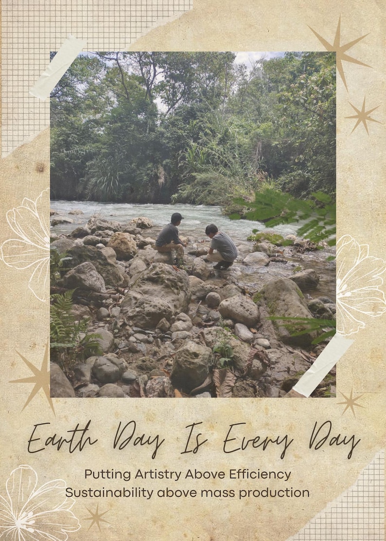 May include: A rectangular graphic print featuring a photograph of two individuals sitting on rocks by a flowing river surrounded by trees. The text reads Earth Day Is Every Day, Putting Artistry Above Efficiency, Sustainability above mass production.