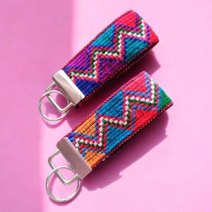 May include: Two colorful woven keychains with a zig-zag pattern in red, blue, purple, and orange. Each keychain has a silver-colored metal clasp and ring. The background is a solid pink.