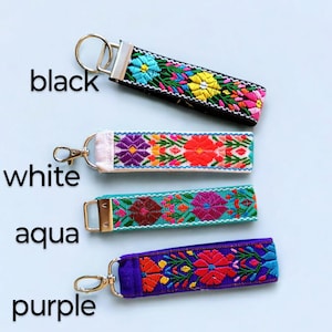 May include: Four colorful embroidered keychains with different colored fabric bases. The keychains are black, white, aqua, and purple. Each keychain has a floral design with bright colors.