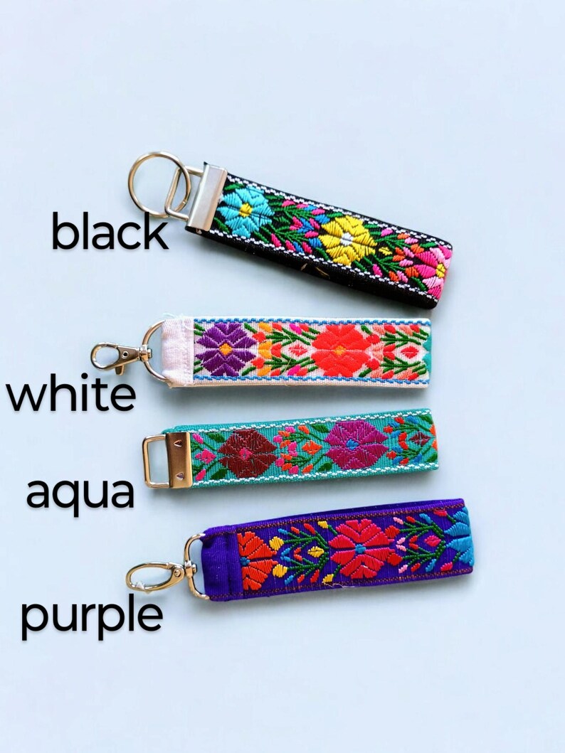 May include: Four colorful embroidered keychains with different colored fabric bases. The keychains are black, white, aqua, and purple. Each keychain has a floral design with bright colors.