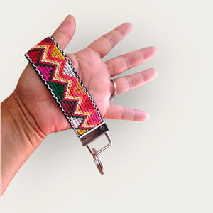 May include: A colorful woven keychain wristlet with a zig-zag pattern in red, pink, yellow, green, and white. The wristlet has a silver-colored metal clasp and key ring. The wristlet is held against a plain background.