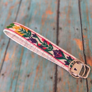 May include: A fabric wristlet keychain featuring a cream-colored base with an embroidered floral pattern in shades of green, yellow, purple, and red. The strap is attached to a silver-toned metal split ring and a rectangular hardware connector.