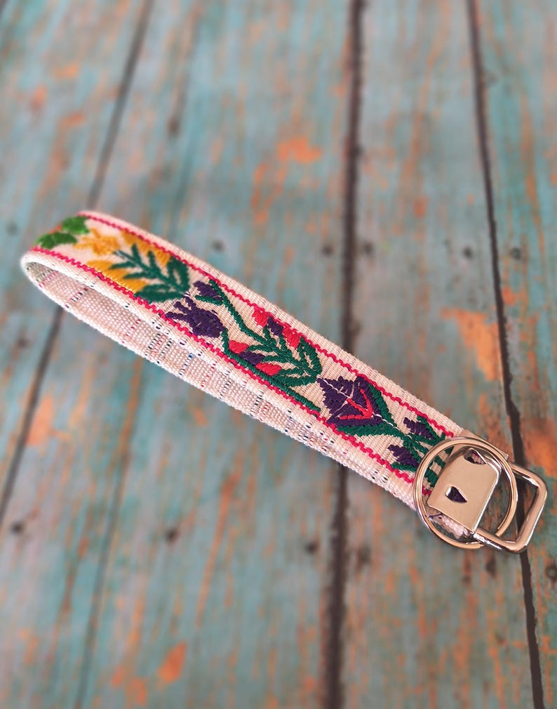 May include: A fabric wristlet keychain featuring a cream-colored base with an embroidered floral pattern in shades of green, yellow, purple, and red. The strap is attached to a silver-toned metal split ring and a rectangular hardware connector.