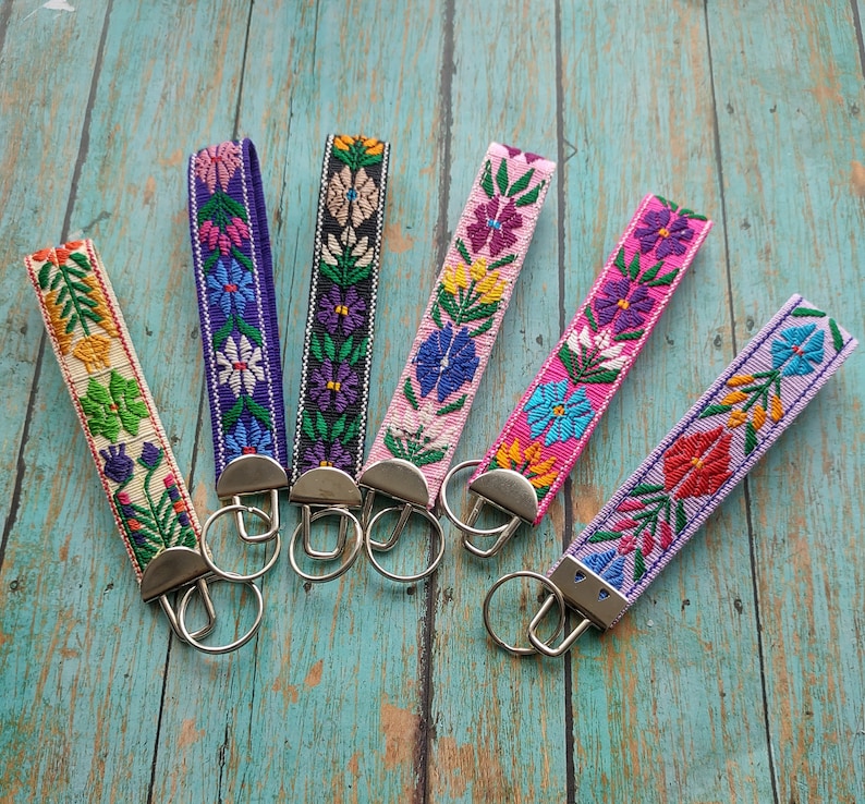 May include: Six fabric keychains featuring colorful floral embroidery patterns on various background colors including cream, purple, black, light pink, hot pink, and lavender, each attached to a silver-toned metal clasp and a circular key ring.