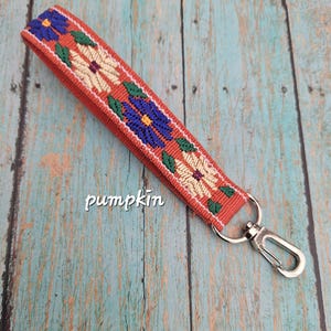 May include: A wristlet keychain featuring a burnt orange fabric strap with an embroidered floral pattern in blue, cream, and green. The strap is attached to a silver-toned metal lobster clasp. The word pumpkin is printed in white text.