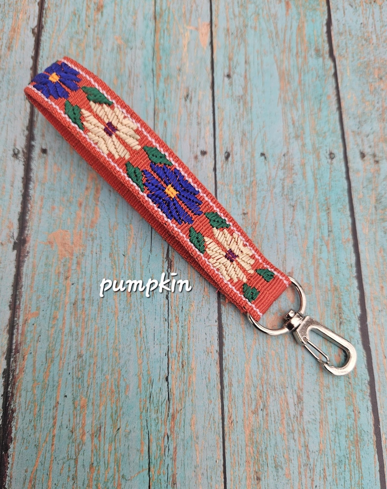 May include: A wristlet keychain featuring a burnt orange fabric strap with an embroidered floral pattern in blue, cream, and green. The strap is attached to a silver-toned metal lobster clasp. The word pumpkin is printed in white text.