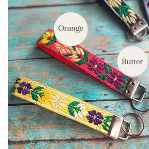 May include: Three fabric keychains featuring embroidered floral patterns in yellow, red, and black. Each strap is finished with a silver-toned metal clamp and a circular split ring. The text labels Orange and Butter are visible on the image.
