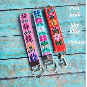 May include: Three fabric wristlet keychains with embroidered floral patterns in pale pink, sky blue, and orange. Each strap features a silver-toned metal clasp and a circular split ring for keys. The text Pale pink, Sky blue, and Orange is visible.
