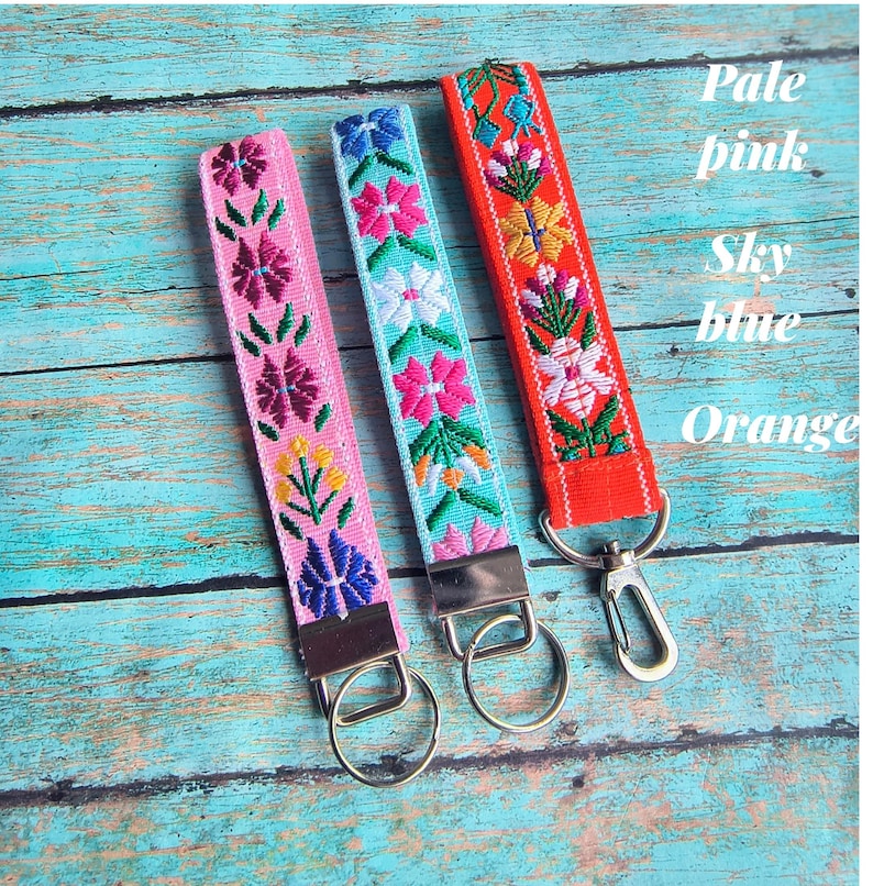 May include: Three fabric wristlet keychains with embroidered floral patterns in pale pink, sky blue, and orange. Each strap features a silver-toned metal clasp and a circular split ring for keys. The text Pale pink, Sky blue, and Orange is visible.