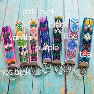May include: Seven fabric keychains with embroidered floral patterns in hot pink, vanilla, pale pink, purple, black, lilac, and teal. Each strap features a silver-toned metal clasp and a circular split ring for keys, arranged in a row.
