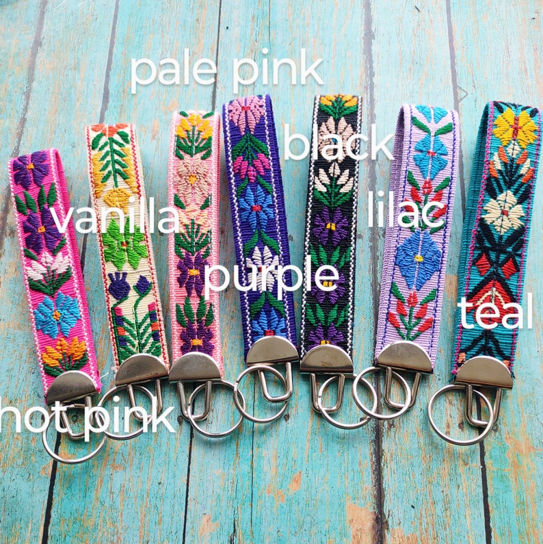 May include: Seven fabric keychains with embroidered floral patterns in hot pink, vanilla, pale pink, purple, black, lilac, and teal. Each strap features a silver-toned metal clasp and a circular split ring for keys, arranged in a row.