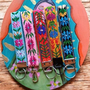 May include: Five fabric wristlet keychains featuring intricate floral embroidery in light blue, pink, red, yellow, and black. Each strap is finished with a silver-toned metal clasp and a circular split ring, except for the yellow one with a black clasp.